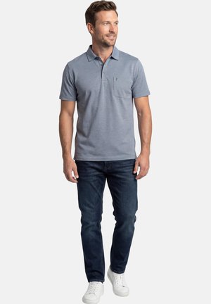 Mężczyzna idący, ubrany w jasnoniebieską krótkorękawową koszulkę polo z kieszenią, ciemnoniebieskie dżinsy i białe trampki, patrzący na bok.