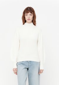 Ung kvinne med rødt hår iført en hvit ribbet turtleneck-genser og lyseblå jeans, stående mot en ensfarget hvit bakgrunn.