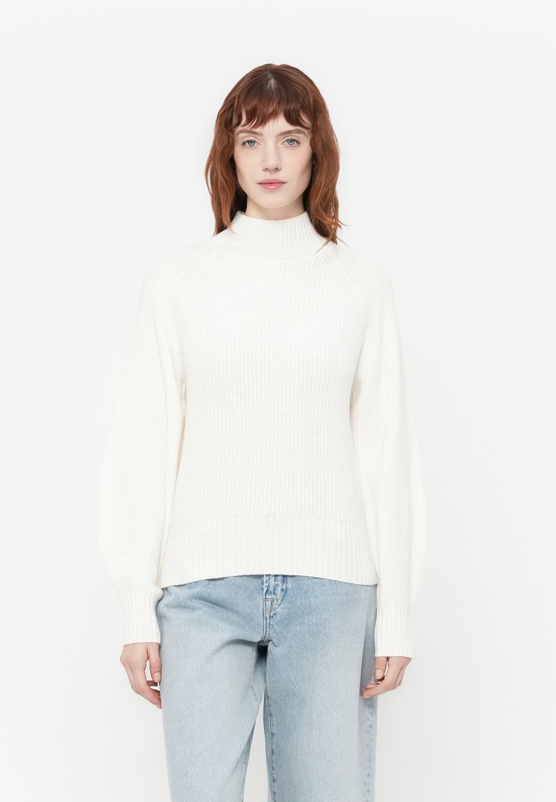 Jeune femme aux cheveux roux portant un pull à col roulé blanc à côtes et un jean bleu clair, se tenant devant un fond blanc uni.