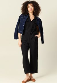 Svart jumpsuit med knytmidja, med korta ärmar och vida ben, kombinerad med en mörk denimjacka. Bruna klacksandaler fulländar looken.