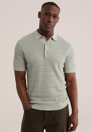 Mann trägt ein hellgrünes, strukturiertes kurzärmeliges Poloshirt und dunkle Hose, steht mit einer Hand in der Tasche vor einem einfarbigen Hintergrund.