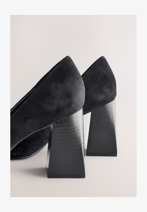 Melni zamšādas augstpapēžu kurpes ar ģeometrisku bloku papēdi. Papēdis ir matēta tekstūra, bet virsma gluda.
