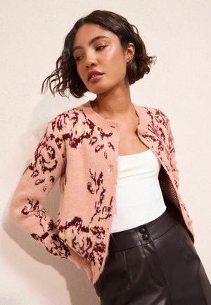 Pletený kardigán svetloružovej farby s vínovými kvetinovými vzormi, okrúhlym výstrihom a zapínaním na gombíky. Kombinovaný s bielym tričkom a tmavými nohavicami.