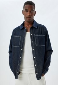 Mörk denimjacka med två framfickor, knappstängning, kontrasterande vit sömnad och en klassisk kragsdesign. Bärs över en vit skjorta.