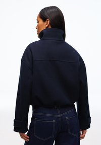 Pitkähiuksinen nainen, jolla on yllään tummansininen kauluksellinen takki ja tummansiniset farkut, selin yksiväristä vaaleaa taustaa vasten.