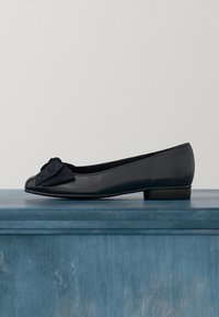 Ballerine en cuir noir avec un petit talon et un grand nœud décoratif à l'avant, exposée sur une surface en bois bleue.