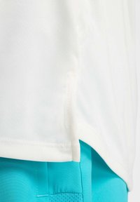 Weißes Sportshirt mit glatter Textur, ausgestattet mit seitlichen Schlitzen am Saum, kombiniert mit leuchtend türkisfarbenen Shorts mit einem Netzfach.