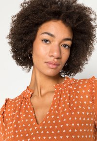 Blusa laranja com pequenos padrões de corações brancos. Apresenta um decote em V e detalhes de ombro com babados. Tecido leve com uma aparência texturizada.
