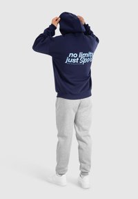 Hoodie marine avec une poche kangourou à l'avant, arborant un texte bleu clair au dos : « pas de limites, juste du Sport ». Associé à un pantalon de survêtement gris et des chaussures blanches.