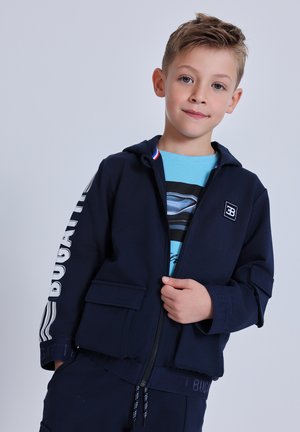 Young boy wearing a navy blue Bugatti zip-up jacket over a blue graphic t-shirt, standing with a slight smile against a plain background.