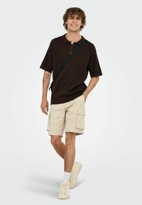 Camisa polo de manga corta marrón con tapeta de tres botones, combinada con pantalones cortos de cargo beige que tienen bolsillos laterales y unas zapatillas de color claro.