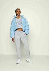 Jonge vrouw in een lichtblauwe cropped pufferjack, grijze cropped sweatshirt, grijze joggingsbroek en witte sneakers, staand tegen een effen achtergrond.