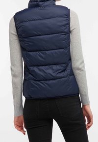 Marineblauw puffergilet met een hoge kraag, horizontaal doorgestikt ontwerp en een gladde textuur. Gedragen over een grijs shirt met lange mouwen, voorzien van zijzakken.