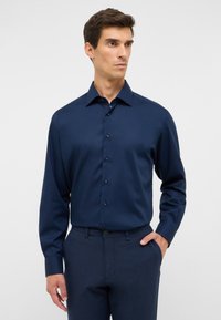 Camicia a maniche lunghe blu navy in tessuto liscio con colletto a punta, chiusura con bottoni e un leggero lucido, abbinata a pantaloni scuri dalla texture. La combinazione offre un look elegante e raffinato.