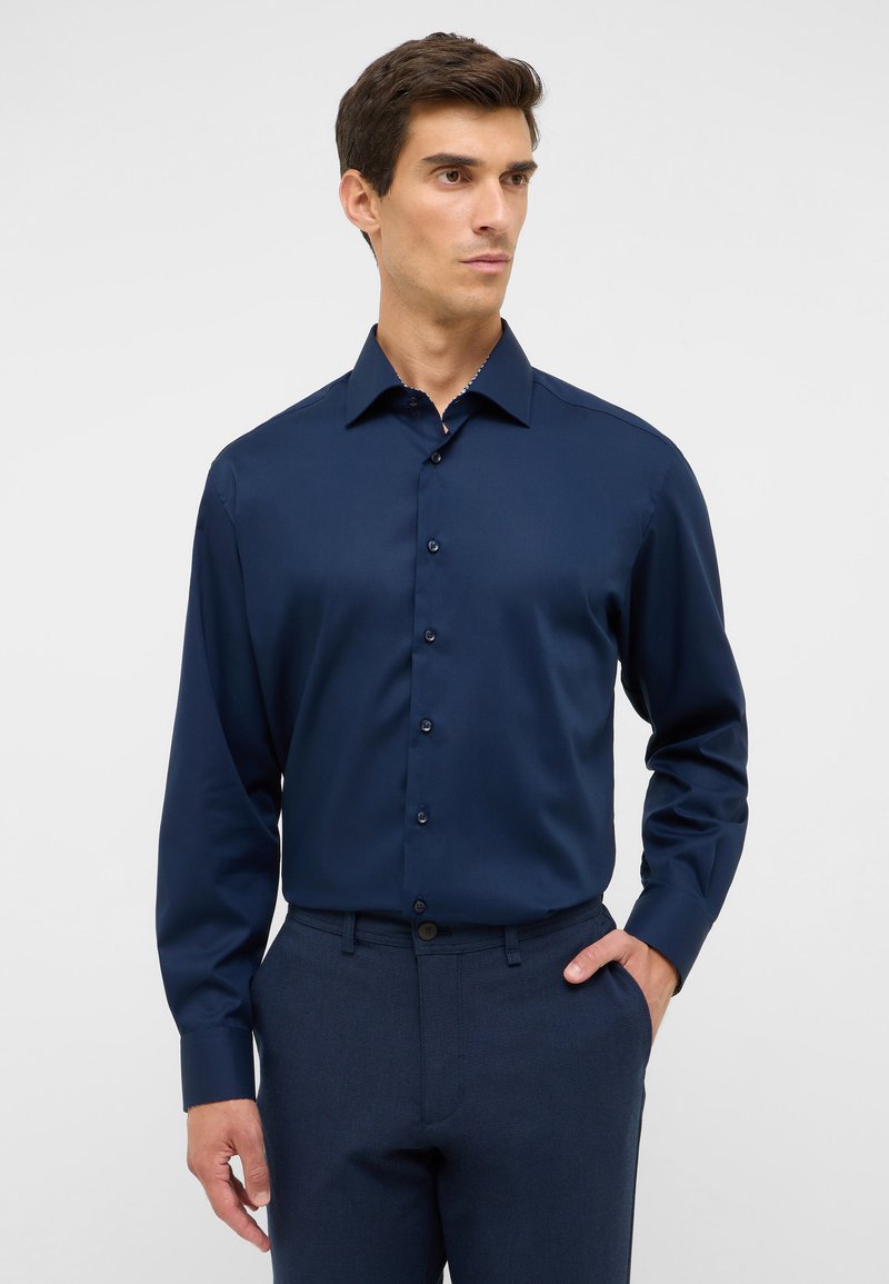 Camicia a maniche lunghe blu navy in tessuto liscio con colletto a punta, chiusura con bottoni e un leggero lucido, abbinata a pantaloni scuri dalla texture. La combinazione offre un look elegante e raffinato.