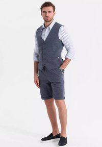 Námořnická modrá vesta přes pruhovanou košili, doplněná ladícími šortkami. Hladká textura, knoflíkové detaily a černé slip-on boty dotvářejí celý outfit.