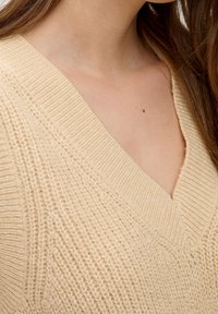 Beige gebreide trui met een V-vormige halslijn. De textuur is ribbelig met zichtbare horizontale lijnen en heeft een relaxte pasvorm en een zachte uitstraling.