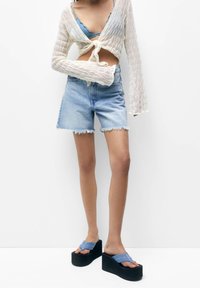 Maglione a maniche lunghe all'uncinetto, beige chiaro; top corto blu; pantaloni corti in denim chiaro con orlo sfrangiato; sandali platform neri con cinturini blu.
