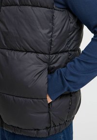 Schwarze, wattierte Weste mit gesteppten Abschnitten, Seitentasche und Reißverschluss. Über einem blauen Langarmshirt mit gerippter Textur getragen.