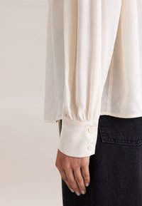 Chemisier blanc en chiffon avec des manches longues, poignets froncés dotés de deux boutons, associé à un jean en denim foncé. Texture douce et design fluide.