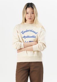 Sweatshirt de culoare crem cu text albastru, guler rotund, mansete și tiv cu margini ondulate. Purtați cu pantaloni maro, cu logo-ul brandului și anul înființării.