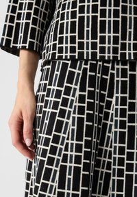 Indumento a motivo geometrico in bianco e nero, caratterizzato da un tessuto testurizzato con forme squadrate e una mano visibile che tiene il tessuto.