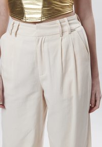 Pantalones de talle alto color crema, hechos de una tela suave, con pliegues y una cinturilla con pasadores para cinturón. Incluye doble bolsillo frontal.