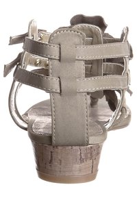 Šedá sandála s blokovým podpatkem, na které je propletený páskový vzor. Materiál působí měkce, s texturovaným dřevěným podpatkem a minimalistickými detaily.