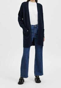 Navy-blue oversized cardigan with open front, long sleeves, and two front pockets, worn over a white shirt and flared denim jeans.
