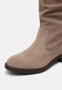 Botte en daim marron clair avec bout arrondi, détails cousus et semelle foncée, vue de profil sur fond blanc.