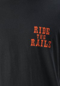 T-shirt de algodão preto com letras laranja em destaque que dizem "CAVALGUE NOS TRILHOS". Textura suave com um design de gola redonda padrão.