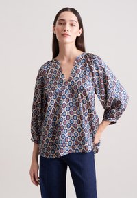 Luftige Bluse aus gemustertem Stoff mit blauem, orangefarbenem und cremefarbenem Blumenmuster, tiefem V-Ausschnitt und Ballonärmeln. Kombiniert mit dunkelblauer Jeans.