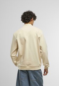 Béžová športová bunda s vysokým golierom a dlhými rukávmi. Vyrobená z mäkkej tkaniny, s rebrovaným lemovaním a bezšvovým dizajnom ramien.