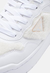 Baskets blanches avec un accent en tissu crème texturé, arborant un logo en triangle doré et des lacets blancs, semelle en caoutchouc et lignes de design épurées.
