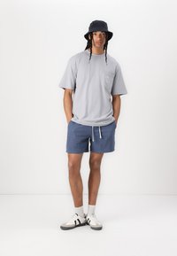 Szary t-shirt z krótkim rękawem i kieszonką, niebieskie spodenki w wzory, czarna kapelusz typu bucket, białe sneakersy z czarnymi paskami i szare skarpetki.