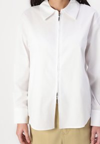 Camicia bianca con colletto a punta, full zip e maniche lunghe. Realizzata in tessuto liscio con un taglio dritto e un leggero dettaglio sull'orlo.