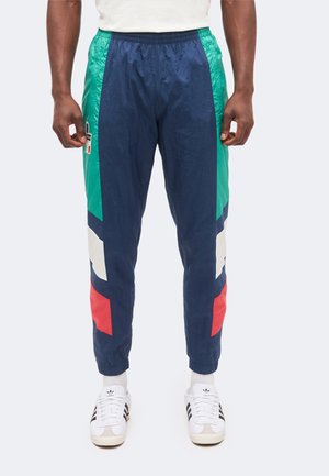 Muž nosi tmavomodré tepláky s farebnými blokmi v zelenej, bielej a červenej farbe, v kombinácii s bielymi teniskami Adidas s čiernymi pruhmi.