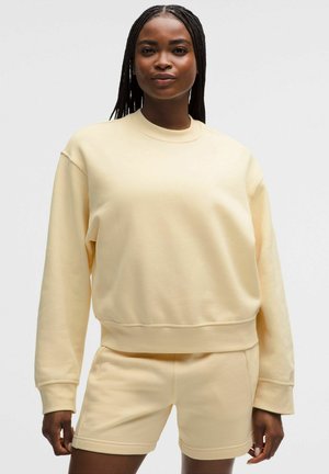 Kvinde med flettet hår iført en lys gul langærmet sweatshirt og matchende shorts, stående mod en ensfarvet lys baggrund.