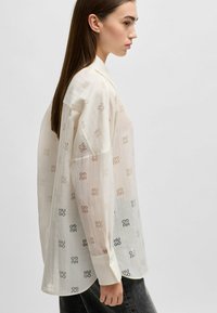 Doorzichtig, crème-kleurig shirt met gepatternde logo's in tinten beige en grijs. Heeft een klassieke kraag en knoopmanchetten. Relaxte pasvorm.