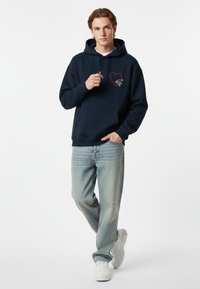 Felpa con cappuccio blu navy, con tasca frontale e grafica a cuore rosso, abbinata a jeans chiari leggermente strappati e sneakers bianche.