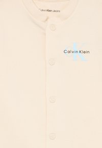 Béžová košeľa s gombíkmi Calvin Klein Jeans, jemná tkanina, okrúhly výstrih, modré a čierne logo na ľavej strane, päť zladených gombíkov vo vertikálnom usporiadaní.