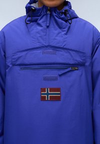 Mėlynas nepralaidus vandeniui džemperis su aukštu apykakle, reguliuojamomis raištelėmis, priekyje užtrauktuku ir šviečiančiu Norvegijos vėliavos ženklu.
