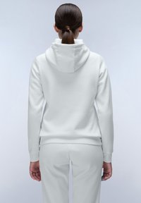 Witte hoodie met een zachte textuur, voorzien van een capuchon en ribgebreide manchetten, gedragen met bijpassende witte broek. zichtbare stikdetails op de achterkant.