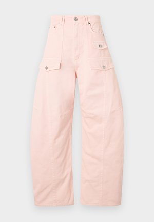 Vysoká žena v bielom tielku bez rukávov, svetloružových širokých cargo nohaviciach, čiernych topánkach a veľkých čiernych slnečných okuliaroch, stojaca s jednou rukou v vrecku.