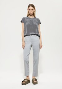 Graue kurzärmelige Bluse mit Faltendetails, kombiniert mit hellgrauen schmalen Hosen und olivgrünen Plattformsandalen. Glatte Texturen überall.