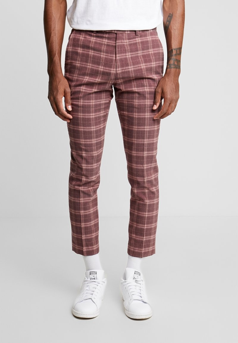 Homme portant un pantalon slim bordeaux à carreaux longueur cheville, des baskets blanches, des chaussettes blanches et un t-shirt blanc, debout sur un sol gris.