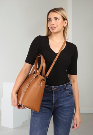 Woman wearing black top and blue jeans holding a tan leather handbag with double straps in a minimalist room.