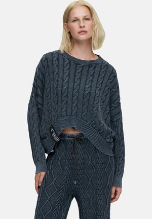 Femme portant un pull torsadé gris foncé et un pantalon en maille texturée assorti avec une taille à cordon, debout devant un fond uni.