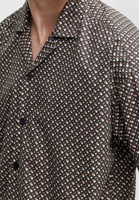 Camisa de manga corta en tejido marrón oscuro con un patrón de cuadros de colores blanco y marrón claro. Presenta un cuello y botones negros.
