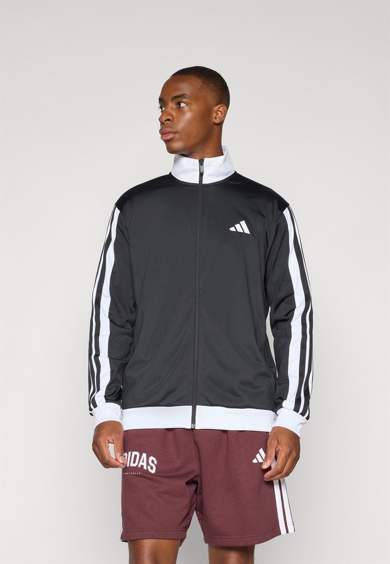 Chaqueta deportiva negra con acentos blancos y tres franjas blancas en cada manga. Está hecha de una tela suave, con un cierre frontal con cremallera y un alto cuello.